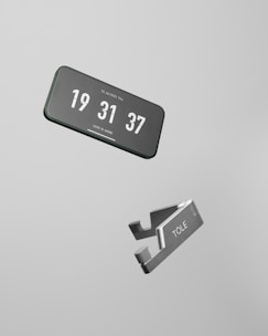 A smartphone displaying the current time floating in mid-air, next to a metallic object labeled 'TOLE'. The background is a plain light gray, emphasizing the minimalist design.