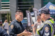 Two police officers are engaged in a conversation in an outdoor setting. The setting appears to be in front of a building with glass windows and is structured with banners and a tent displaying police logos. Other individuals are seen in the background, indicating a public or community event.