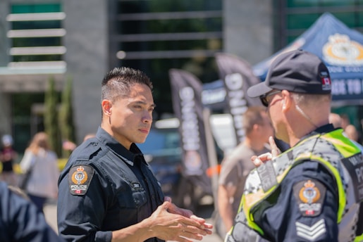 Two police officers are engaged in a conversation in an outdoor setting. The setting appears to be in front of a building with glass windows and is structured with banners and a tent displaying police logos. Other individuals are seen in the background, indicating a public or community event.