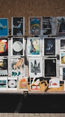 A collection of various posters and artworks is displayed on a wooden board. The images vary in style and content, including abstract art, portraits, and graphic designs. Some feature text in an unknown language, while others depict illustrations, paintings, or digitally altered photos. The board is set in an outdoor location, with sunlight casting shadows across the images.