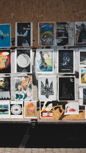 A collection of various posters and artworks is displayed on a wooden board. The images vary in style and content, including abstract art, portraits, and graphic designs. Some feature text in an unknown language, while others depict illustrations, paintings, or digitally altered photos. The board is set in an outdoor location, with sunlight casting shadows across the images.