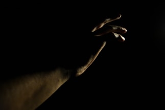 A softly lit, sculptural hand reaching out against a dark satin-matte background with a warm yellow accent.
