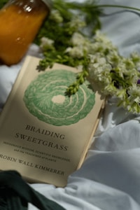 A book titled 'Braiding Sweetgrass' by Robin Wall Kimmerer is placed on a white fabric, surrounded by white flowers and a glass jar filled with an orange liquid. The book cover features a green braided design.