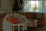 A cozy living room features a hanging macrame chair with an orange cushion and fringed throw. In the background, a large brown leather sofa is visible near a wooden coffee table topped with a plant. Sheer curtains allow natural light to filter through a window, creating a warm and inviting atmosphere.