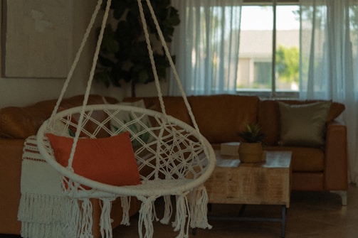 A cozy living room features a hanging macrame chair with an orange cushion and fringed throw. In the background, a large brown leather sofa is visible near a wooden coffee table topped with a plant. Sheer curtains allow natural light to filter through a window, creating a warm and inviting atmosphere.