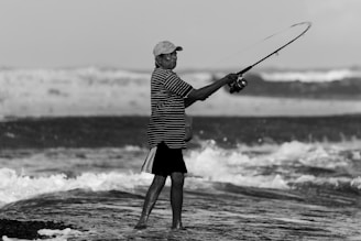 A person is standing in shallow ocean water holding a fishing rod, casting a line into the waves. The scene is captured in black and white, highlighting the contrast of the waves against the shoreline. The individual wears a striped shirt, shorts, and a cap, conveying a casual and relaxed atmosphere in this natural setting.