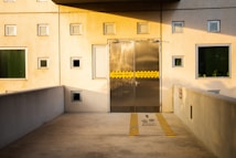 A modern building facade featuring a wall with multiple square windows and a metal door in the center. The door is marked with yellow caution tape indicating it opens from the inside. The wall and surrounding areas are illuminated by the warm light of the setting sun, casting shadows and creating a serene atmosphere. The pathway leading to the door has yellow and black striped markings with a cautionary message and directional arrows pointing straight ahead.