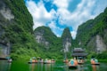 Tourists in orange life jackets are paddling boats through a serene river surrounded by towering limestone mountains covered in lush green vegetation. A traditional Asian pagoda stands in the middle of the river, enhancing the picturesque landscape.