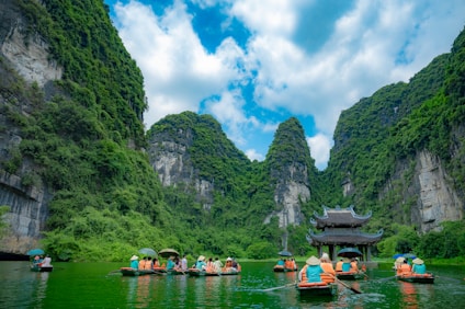 Tourists in orange life jackets are paddling boats through a serene river surrounded by towering limestone mountains covered in lush green vegetation. A traditional Asian pagoda stands in the middle of the river, enhancing the picturesque landscape.