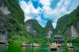 Tourists in orange life jackets are paddling boats through a serene river surrounded by towering limestone mountains covered in lush green vegetation. A traditional Asian pagoda stands in the middle of the river, enhancing the picturesque landscape.
