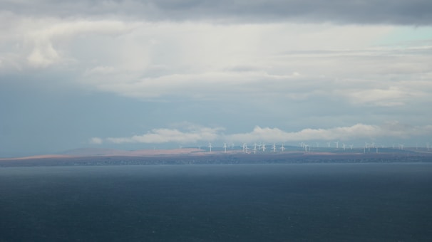 A vast body of water extends across the foreground with a distant shoreline visible in the background. On the horizon, numerous wind turbines are spread across the landscape under a cloudy sky, suggesting renewable energy use.