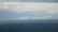 A vast body of water extends across the foreground with a distant shoreline visible in the background. On the horizon, numerous wind turbines are spread across the landscape under a cloudy sky, suggesting renewable energy use.