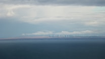 A vast body of water extends across the foreground with a distant shoreline visible in the background. On the horizon, numerous wind turbines are spread across the landscape under a cloudy sky, suggesting renewable energy use.
