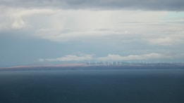 A vast body of water extends across the foreground with a distant shoreline visible in the background. On the horizon, numerous wind turbines are spread across the landscape under a cloudy sky, suggesting renewable energy use.