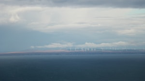 A vast body of water extends across the foreground with a distant shoreline visible in the background. On the horizon, numerous wind turbines are spread across the landscape under a cloudy sky, suggesting renewable energy use.