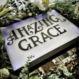 A decorative sign surrounded by lush greenery and white flowers prominently displays the words 'Amazing Grace' in an ornate, stylized font. The arrangement includes various types of leaves and blooms, creating a vibrant and serene atmosphere.