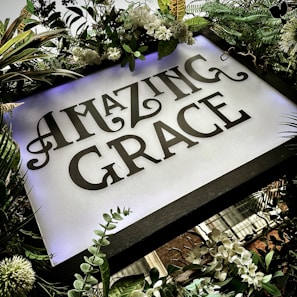 A decorative sign surrounded by lush greenery and white flowers prominently displays the words 'Amazing Grace' in an ornate, stylized font. The arrangement includes various types of leaves and blooms, creating a vibrant and serene atmosphere.