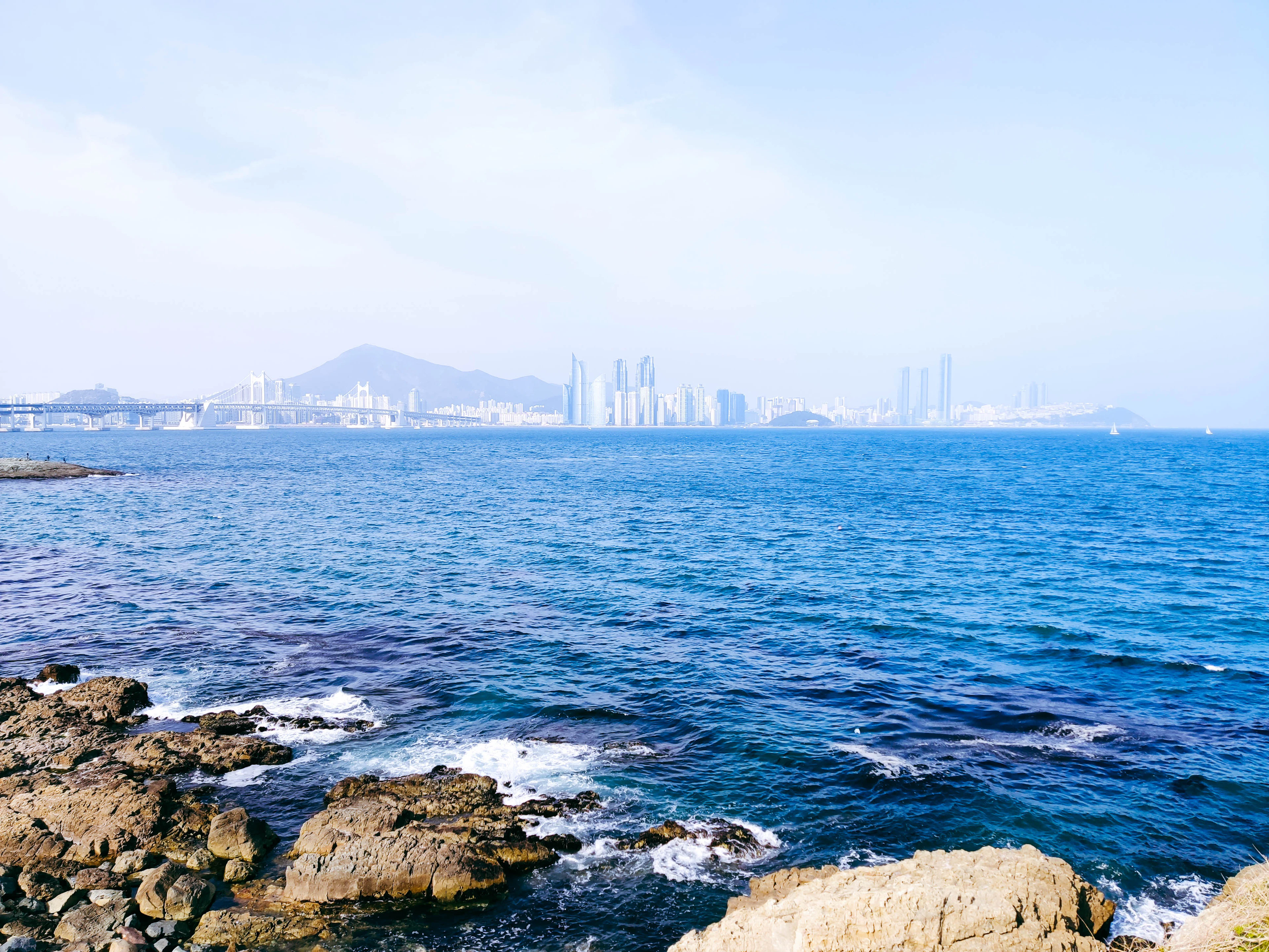 Rocky coastline at Igidae with the high-rise Haeundae skyline across blue water.