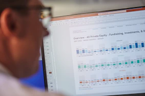 A person is looking at a computer screen displaying a document titled 'Overview - All Private Equity - Fundraising, Investments, & D'. The document contains statistical charts and graphs related to private equity market statistics from 2007 to 2022. Various tabs and functions like 'File', 'Home', 'Insert', and 'Design' are visible in the interface.