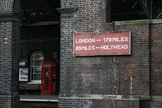 A vintage railway station scene with a prominent brick wall displaying a directional sign for London and Holyhead. A classic red British telephone box is visible next to an arched window and a station bench.