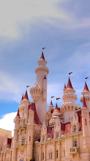A grand fairy-tale castle with multiple tall spires and pointed red roofs topped with small flags. The castle is bathed in bright sunlight against a backdrop of a clear blue sky with some clouds.
