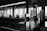 A series of abstract geometric sculptures stand in the foreground of a modern, minimalistic interior space. The sculptures consist of stacked rectangular prisms, creating a city-like skyline. In the background, large floor-to-ceiling windows reveal blurred cityscape views, while silhouettes of people are seated, contributing to a contemplative atmosphere.