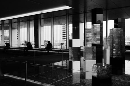 A series of abstract geometric sculptures stand in the foreground of a modern, minimalistic interior space. The sculptures consist of stacked rectangular prisms, creating a city-like skyline. In the background, large floor-to-ceiling windows reveal blurred cityscape views, while silhouettes of people are seated, contributing to a contemplative atmosphere.