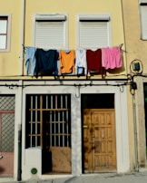 Laundry, including shirts and towels, is hanging on a clothesline across an upper floor of a building with a pale yellow facade. Below, there are two closed windows with shutters, a wooden door, and barred windows. The overall setting suggests a residential area in an urban environment.