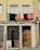 Laundry, including shirts and towels, is hanging on a clothesline across an upper floor of a building with a pale yellow facade. Below, there are two closed windows with shutters, a wooden door, and barred windows. The overall setting suggests a residential area in an urban environment.