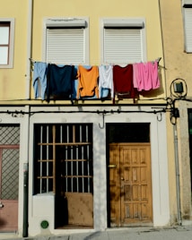 Laundry, including shirts and towels, is hanging on a clothesline across an upper floor of a building with a pale yellow facade. Below, there are two closed windows with shutters, a wooden door, and barred windows. The overall setting suggests a residential area in an urban environment.