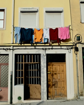 Laundry, including shirts and towels, is hanging on a clothesline across an upper floor of a building with a pale yellow facade. Below, there are two closed windows with shutters, a wooden door, and barred windows. The overall setting suggests a residential area in an urban environment.