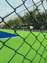 A sports court with vibrant green and blue turf is viewed through a chain-link fence. In the background, a few trees and vehicles are visible. The sky is partly cloudy, and a yellow pole is situated on the court.