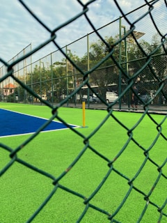 A sports court with vibrant green and blue turf is viewed through a chain-link fence. In the background, a few trees and vehicles are visible. The sky is partly cloudy, and a yellow pole is situated on the court.
