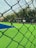 A sports court with vibrant green and blue turf is viewed through a chain-link fence. In the background, a few trees and vehicles are visible. The sky is partly cloudy, and a yellow pole is situated on the court.