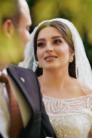 A bride wearing an elegant, beaded white wedding dress gazes lovingly at someone in a black suit. Her makeup is elaborate, and she wears a pearl-covered veil.