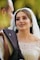 A bride wearing an elegant, beaded white wedding dress gazes lovingly at someone in a black suit. Her makeup is elaborate, and she wears a pearl-covered veil.