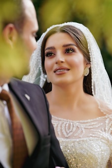 A bride wearing an elegant, beaded white wedding dress gazes lovingly at someone in a black suit. Her makeup is elaborate, and she wears a pearl-covered veil.