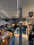 The interior of a small convenience store filled with various packaged goods such as drinks, snacks, and groceries. The shelves are neatly organized with many products on display, and signs are hanging from the ceiling advertising promotions. Two people are present; one is standing near the counter at the back making a transaction while the other is browsing or reaching for an item on a lower shelf.