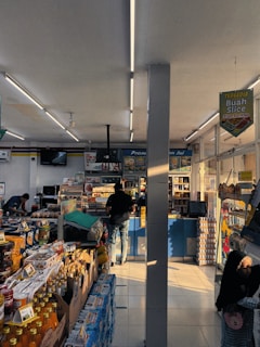 The interior of a small convenience store filled with various packaged goods such as drinks, snacks, and groceries. The shelves are neatly organized with many products on display, and signs are hanging from the ceiling advertising promotions. Two people are present; one is standing near the counter at the back making a transaction while the other is browsing or reaching for an item on a lower shelf.