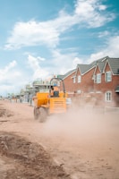 A construction vehicle is driving through a dusty path flanked by newly built brick houses and construction scaffolding. The clear blue sky with scattered clouds adds contrast to the scene. The vehicle is bright orange, stirring up a significant amount of dust as it moves along the dirt road.