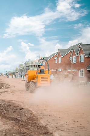 A construction vehicle is driving through a dusty path flanked by newly built brick houses and construction scaffolding. The clear blue sky with scattered clouds adds contrast to the scene. The vehicle is bright orange, stirring up a significant amount of dust as it moves along the dirt road.
