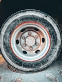 A close-up view of a weathered truck tire mounted on a steel wheel with visible rust and dirt buildup. The circular arrangement of bolt holes and a central hub are prominent. The tire appears to be slightly worn and rests on a textured concrete surface.