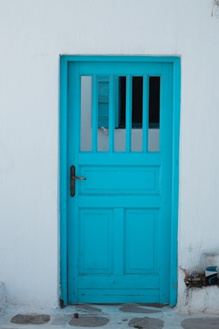 A vibrant turquoise wooden door set in a white plaster wall, with a metal handle on the left side. The upper part of the door features vertical rectangular glass panels allowing some light to pass through. The ground outside is covered with stone tiles.