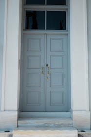 A tall, elegant double door painted in light blue with six rectangular panels on each side. The door is framed by a simple white structure and features gold handles and a keyhole. Above the door, there are three rectangular transom windows reflecting a dark, outdoor silhouette.