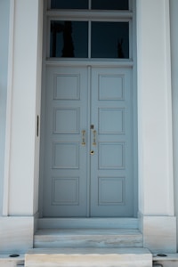 A tall, elegant double door painted in light blue with six rectangular panels on each side. The door is framed by a simple white structure and features gold handles and a keyhole. Above the door, there are three rectangular transom windows reflecting a dark, outdoor silhouette.