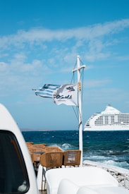 A large cruise ship sails on a deep blue sea under a partly cloudy sky. In the foreground, several wicker chairs are arranged, and two flags are fluttering in the breeze, one of them being the Greek flag.