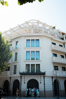 A multi-story building with a decorative modern facade and large windows. There are arched entrances at the ground level and an open terrace with a geometric framework at the top. People stand around a display of informational brochures near the building.