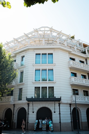 A multi-story building with a decorative modern facade and large windows. There are arched entrances at the ground level and an open terrace with a geometric framework at the top. People stand around a display of informational brochures near the building.
