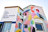 A colorful building facade with abstract mural art and a motivational phrase on the left reading 'eat. sleep. sun. après-beach. repeat.' The art includes shapes in various colors including pastels and bright hues, with a mix of geometric and fluid designs. The windows are integrated into the mural, adding depth and interest to the artwork.