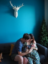 A couple is sitting on a dark grey couch, tenderly holding a baby. They are wearing matching avocado-patterned clothing. A 3D geometric white deer head is mounted on a bright teal wall. A small evergreen tree stands in the corner. A plush toy is partially visible on the couch.
